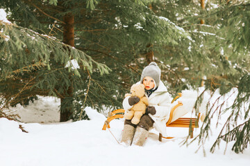 cute little boy toddler in fur coat, knitted hat and felt boots sitting on sled with teddy bear toy in snow winter forest, concept of Christmas holidays and New Year in retro vintage style