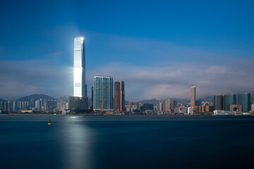 Fototapeta na wymiar Lighthouse. The sun reflects off a skyscraper on Kowloon, Hong Kong casting light over the blue sea. Long exposure gives a silky smooth sea.