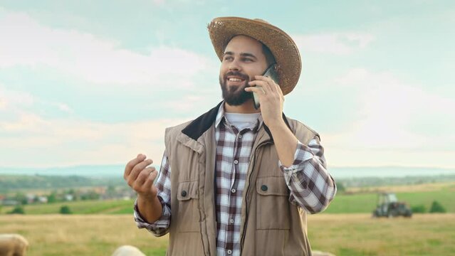 Caucasian handsome man in hat standing in grassland and talking on cellphone. Outside. Good-looking young male shepherd speaking and chatting on mobile phone. Sheep grazing. Countryside concept.