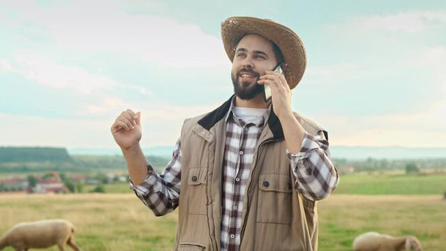 Caucasian attractive man in hat standing in field and talking on telephone. Outdoor. Happy handsome young male shepherd speaking and chatting on cellphone. Sheep feeding. Countryside concept.