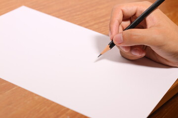 a hand is holding pencil and writing on white paper which is on a wooden table, for placing text or using as background