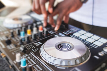 View of Dj mixer and vinyl plate with headphones on a table with african american DJ playing on stage and mixes the track in the background, during summer open air event techno party, hand close up - Powered by Adobe