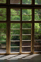 Interior of cottage house in process of renovation in countryside on sunny summer day. Sunlight breaks through big wooden frame of panoramic window overlooking garden with lush green plants and bushes