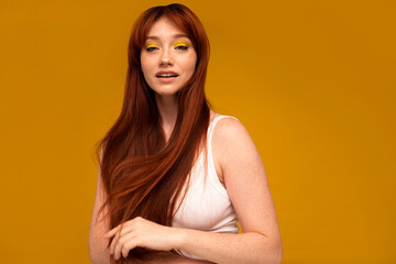 Obraz premium Positive ginger girl with yellow makeup, natural freckles, long hair and fringe. Happy red head woman posing in studio, looking at the camera.