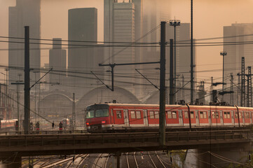 Ein städtischer Nahverkehrszug fährt im Morgendunst oder Abendlicht vor der im Nebel stehenden Skyline der Hochäuser von Frankfurt am Main - obrazy, fototapety, plakaty