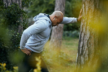 African American sportsman leaning on tree after running in autumn park.