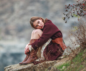 Young lady in long dress and brown boots sitting on stones, on background of rocks and lake in autumn countryside, fashion stylish woman