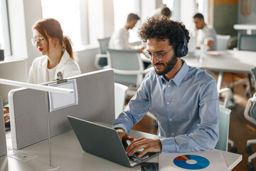 Obraz premium Businessman in headphones working laptop while sitting in modern office on colleagues background