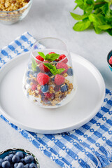 Glass with granola, yogurt, fresh raspberries, blueberries and mint. High protein and diet breakfast on a white-blue napkin. Healthy food concept