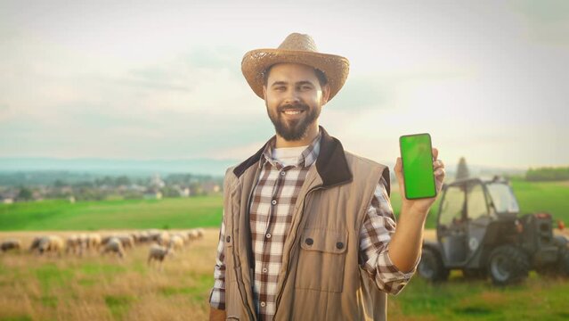 Portrait of handsome young man farmer in hat smiling to camera and showing smartphone with green screen. Chroma key. Attractive male shepherd holding cellphone and demonstrating ads. Sheep and tractor