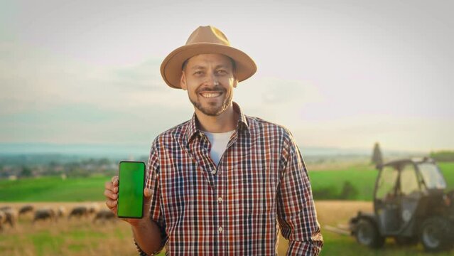Portrait of cheerful young man farmer in hat smiling to camera and showing smartphone with green screen. Chroma key. Attractive happy male shepherd holding cellphone and demonstrating advertisement.