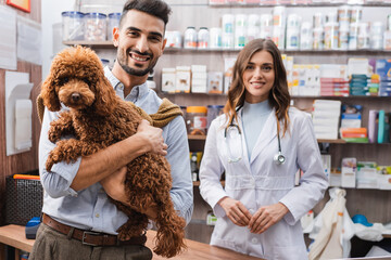 Smiling arbian man holding poodle near veterinarian in pet shop