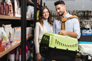 Smiling multiethnic couple looking at animal cage near showcase in pet shop