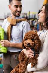 Blurred woman holding brown poodle near smiling muslim boyfriend in pet shop