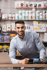 Cheerful arabian seller looking at camera near laptop and payment terminal in pet shop
