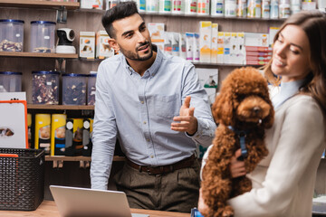 Muslim salesman pointing at blurred woman with poodle in pet shop