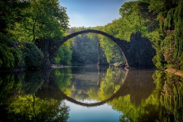 Beautiful view of an arched bridge in the forest symmetrically reflecting on the river water