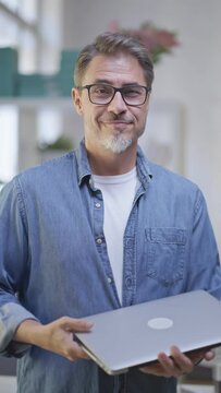 Portrait of middle aged man standing k in bright room working with laptop computer in home office. Mature age, middle age, mid adult casual man in 50s, confident happy smiling.