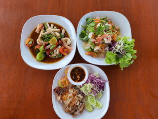 Top view of spicy food consists of salad flow lotus (root lotus) with boiled shrimp, grilled chicken with Thai spicy sauce in a cup, Seafood Glass Noodle Salad Served in a white plate on a wooden tabl