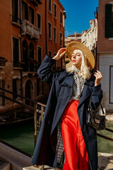 Fototapeta na wymiar Elegant fashionable blonde woman wearing straw hat, classic blue trench coat, polka dot blouse, red skirt, holding handbag, posing on the bridge in Venice. Fashion, travel, lifestyle conception