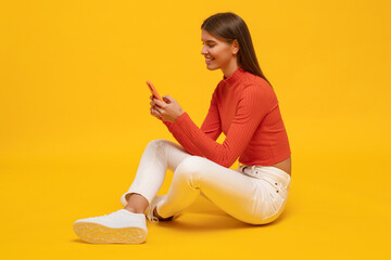 Side view portrait of woman with mobile phone sitting on floor isolated on yellow background