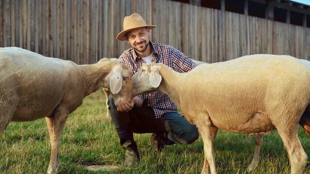 Portrait of cheerful handsome young man shepherd playing with flock of lambs and feeding animals in farm. Outdoor. Happy joyful male farmer in motley shirt and hat smiling to camera at herd of sheep.