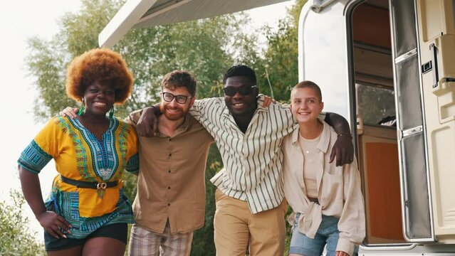 Interracial friendship on a camping van trip. Diverse people, four close friends hugging, smiling, showing a thumb up and a peace sign, taking photos together near their temporary house on wheels