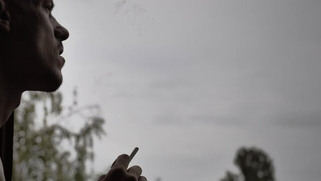 A cinematic dramatic scene, the silhouette of a man who lights a cigarette and smokes slowly releasing smoke. Backlit pensive man in the backlight is sad against the background of a cloudy sky.