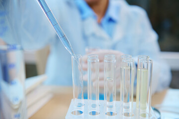 Focus on a glass pipette with a dripping reagent in a test tube on tripod, over blurred background of a chemical laboratory assistant conducting clinical analysis in the biological chemical laboratory
