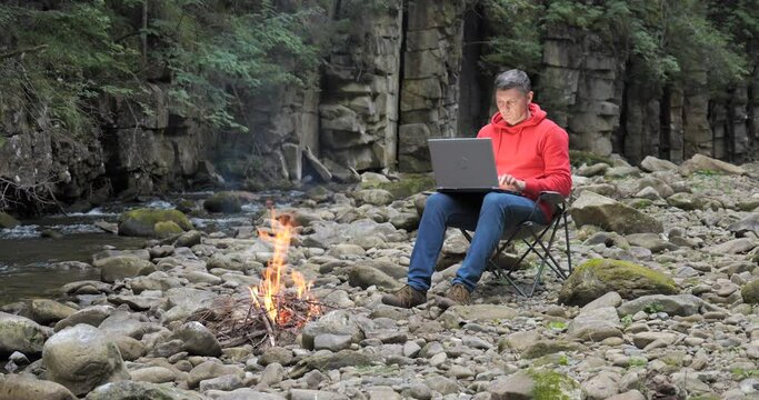 A man works on a laptop near a stream in nature