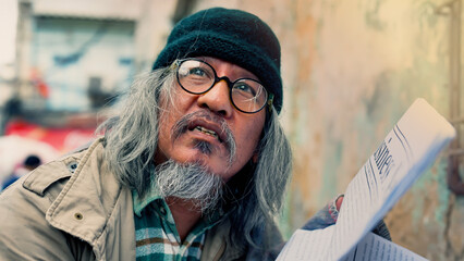 An old Asian man in glasses and a black hat, dressed in a dirty, homeless man, is reading the...
