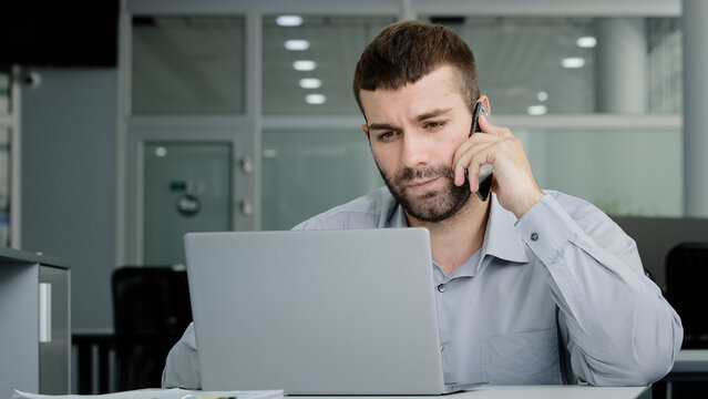 Concentrated young businessman speaks on phone consults client checks data on laptop screen serious manager remote seller offers services over telephone discussing online project multitasking concept