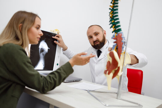 Adult male orthopedic doctor or traumatologist tells the young woman patient about her diagnosis on the model of the spine and x-ray images