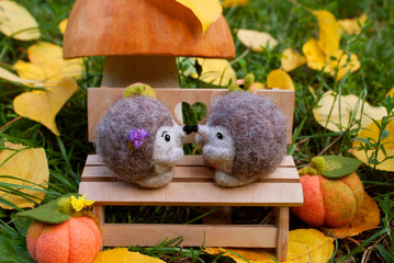 Date under a mushroom made of pumpkin and salt dough, on a plywood bench near two felted hedgehogs during the day, surrounded by green grass, felted pumpkins and yellow leaves