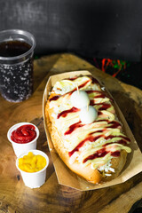 Fast street food, hot dog with sauces, cheese and two quail eggs on top served on a wooden tray and next to it there are two plastic cups with ketchup and mustard, next to the black soda.