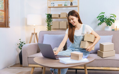 Online merchant concept, Female entrepreneur holds parcel boxes and checks online orders on laptop