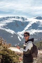 Handsome caucasian man posing to camera. He carries his cell phone and sunglasses. he has snowy mountains in the background