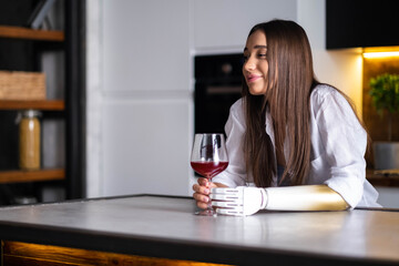 Happy girl with prosthetic arm holds glass of wine in artificial prosthetic limb hand at home