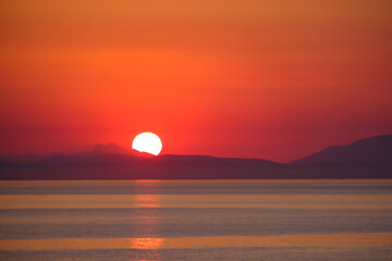 View of the sun rising behind the mountains on the horizon. The sea is in the foreground.