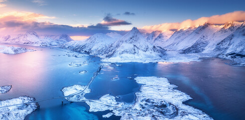 Aerial view of sea, snowy islands, mountains, road, sky with pink clouds at sunset in winter. Lofoten islands, Norway. Landscape with rocks in snow, water at dusk. Top view from above. Nature