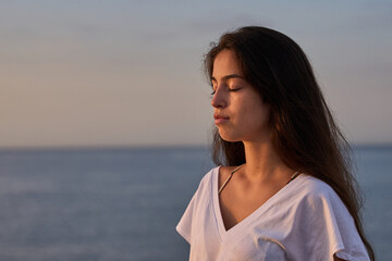 young hispanic tanned girl with a white t-shirt meditating at the rocks early in the morning in the seaside