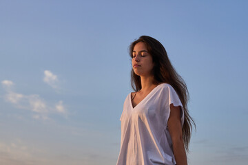 young hispanic tanned girl with a white t-shirt meditating at the rocks early in the morning in the seaside