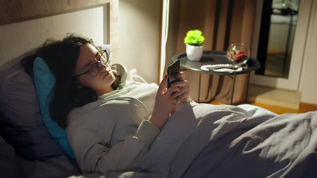 Teenage girl in glasses lying in bed at night with a cell phone in her hands, light from a lamp, camera tracking