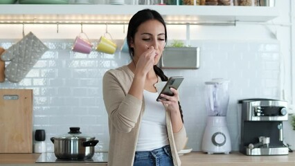 Video of pretty woman sending messages with mobile phone while eating yogurt int he kitchen at home. - Powered by Adobe