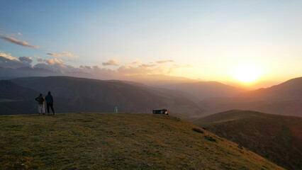 Tourists in the mountains watching sunset. An epic yellow-red sunset is reflected on small clouds and green hills. Bushes and forest grow in places on mountains. People are standing with their backs