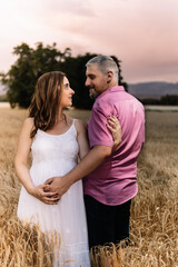 Young and beautiful pregnant couple walking through a wheat field at sunset, with a look of complicity. Concept of happy family.