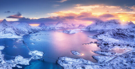 Aerial view of blue sea, snowy islands, mountains, sky with pink clouds at sunset in winter. Lofoten islands, Norway. Landscape with rocks in snow, reflection in water. Top view from drone. Nature