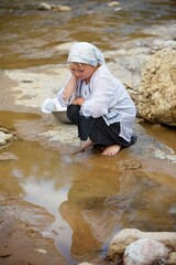 A girl in a long village vintage clothes sitting on the Bank of the river and looking into the water. The river with a fast current in the country with a stone Bank.