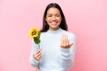 Young Colombian woman holding sunflower isolated on pink background inviting to come with hand. Happy that you came