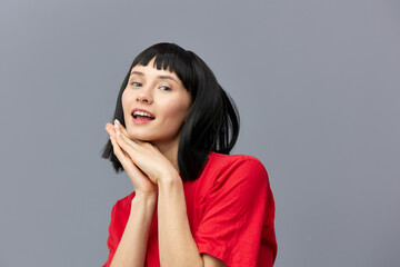 Fototapeta premium a sweet, gentle, sophisticated, relaxed woman with black hair stands in a red T-shirt on a gray background and holds her hands near her face
